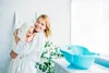 A mother is holding her baby, who is wrapped in a towel, in a bathroom setting. A blue baby bathtub is visible on a counter. Produktbild-Vorschau