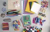 A flat lay of various craft supplies, including paper, pipe cleaners, feathers, pom-poms, glitter, and wooden craft sticks. Produktbild-Vorschau