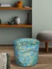 A round ottoman with a floral print is placed on a wooden floor next to a rug. Shelves with decorative items are visible in the background. Produktbild-Vorschau