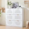 A white shoe cabinet with six flip-down doors, decorated with arched and rectangular panels featuring vertical lines. It is placed in a well-lit room with a wooden floor and a wall shelf above. Produktbild-Vorschau