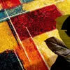 A close-up of a colorful abstract patterned rug with large geometric shapes in red, yellow, orange, and blue. Two large dark green leaves are in the foreground, partially obscuring the rug. Produktbild-Vorschau
