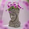A bronze-colored planter shaped like a woman's head, filled with purple petunias, sits on a white surface against a blurred pink and white background. Náhled obrázku produktu