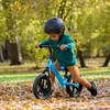 A young child is riding a blue balance bike in an outdoor park setting with fallen leaves on the ground. Produktbild-Vorschau