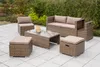 A rattan garden furniture set with cushions and a glass-topped coffee table is arranged on a paved patio, with greenery in the background. Produktbild-Vorschau