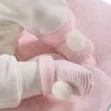A close-up shot of a baby's feet and legs wrapped in pink knitted booties with white pom-poms, resting on a pink knitted blanket. Podgląd zdjęcia produktu