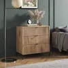 A wooden dresser with three drawers is placed in a room with a dark green wall and a sofa in the background. Produktbild-Vorschau