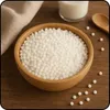 A wooden bowl filled with tapioca pearls on a wooden surface, with a glass of milk and a wooden spoon in the background. Produktbild-Vorschau