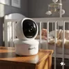 A baby monitor camera is placed on a wooden table in front of a crib where a baby is sleeping. Náhled obrázku produktu