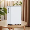 A white dehumidifier with black accents sits on a wooden table. Green plants and beige curtains are visible in the background. Produktbild-Vorschau