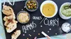 A spread of food items including olives, hummus, and curry dip on a black slate board, with bread slices and confetti scattered around. Produktbild-Vorschau