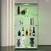 A white glass display cabinet with four shelves, illuminated by green LED lights, showcasing various items including bottles, books, and decorative figures. Podgląd zdjęcia produktu