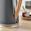 A close-up of a kettle with a wooden handle and a blue indicator light, placed on a wooden surface. A jar of jam and some toast are visible in the background. Podgląd zdjęcia produktu