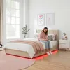 A young woman is sitting on a bed in a bedroom, looking at her phone. The bed has a beige upholstered frame and a red LED light strip around the base. There are pillows and a pink throw blanket on the bed. A nightstand with a lamp is next to the bed. A lar Produktbild-Vorschau