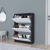 A dark gray shoe cabinet with three white flip-down compartments, each containing shoes, is shown against a light gray wall. A circular mirror hangs on the wall to the left, and a stool is partially visible in the foreground. Náhled obrázku produktu