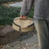 A person is holding a round, tan bag with a black zipper outdoors. Náhled obrázku produktu