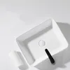 A white ceramic washbasin with a black drain stopper and a small white rolled towel next to it, set against a white background. Podgląd zdjęcia produktu
