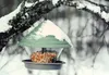 A bird feeder filled with seeds hangs from a snow-covered branch in a winter setting. Produktbild-Vorschau