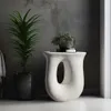 A modern white side table with a unique sculptural design, placed next to a tall potted plant against a textured gray wall. Náhled obrázku produktu