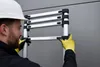 A person wearing a hard hat and yellow gloves is adjusting an aluminum telescopic ladder. Náhľad obrázku produktu