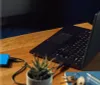 A black laptop is connected to a blue external hard drive on a wooden desk. A small potted plant and keys are also visible. Produktbild-Vorschau