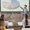 A man in a suit is giving a presentation to an audience, pointing at a pie chart on a projector screen. Produktbild-Vorschau