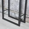 A close-up view of the black metal frame of a firewood rack against a textured wall and light wooden floor. Podgląd zdjęcia produktu
