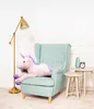 A plush unicorn toy is lying on a light blue armchair, next to a floor lamp and two small tables with decorative items. Podgląd zdjęcia produktu