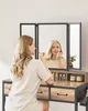 A woman is applying makeup in front of a tri-fold mirror on a vanity. Náhľad obrázku produktu