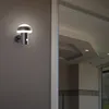 An outdoor wall lamp is mounted on a textured gray wall next to a doorway, with a reflection of an interior ceiling in the glass door. Produktbild-Vorschau