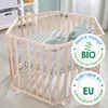 A hexagonal wooden playpen with a grey patterned mat and a green star-shaped pillow inside, set in a room with a white shelf in the background. Produktbild-Vorschau