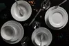 A flat lay of white porcelain dinnerware set on a black surface, with cutlery and glasses. Náhľad obrázku produktu