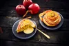 Two apple pastries are presented on dark plates, with whole apples and cinnamon sticks in the background, set against a dark wooden surface. Produktbild-Vorschau