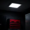 A square LED panel light is mounted on a dark ceiling above a red and black tool chest in a dimly lit room. Náhľad obrázku produktu