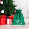 A green Christmas gift bag with white snowflakes and a Santa Claus illustration sits on a wooden floor next to a decorated Christmas tree and wrapped presents. Produktbild-Vorschau