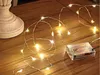 A string of battery-operated LED lights is spread across a wooden surface, with a white decorative base on the left and a clear battery pack on the right. Náhled obrázku produktu
