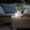 An outdoor patio setting at dusk, featuring a wicker coffee table with a portable LED lamp, a vase of flowers, and candles. A sofa is visible in the background. Anteprima dell’immagine prodotto