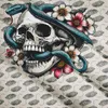 A detailed illustration of a skull entwined with a snake and surrounded by flowers, set against a patterned background. Náhled obrázku produktu