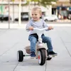 A young child is riding a beige and white tricycle outdoors on a paved walkway. Produktbild-Vorschau