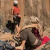 Two people are rock climbing. One is climbing the rock face, and the other is sitting on the ground, preparing to climb. Produktbild-Vorschau