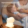 A person is feeding a baby with a bottle, with a dinosaur-shaped night light on a table in the foreground. Voir les images du produit
