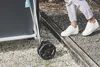 A person's legs and feet wearing white sneakers are shown stepping over a wooden beam next to a blue and gray outdoor table tennis table with black wheels. Produktbild-Vorschau
