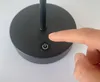A finger is pressing a power button on the base of a black lamp. Produktbild-Vorschau