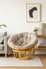 A papasan chair with a cushion is placed on a rug in a living room, next to a sofa and a shelf with a lamp and flowers. Produktbild-Vorschau