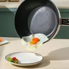 Schwarze Antihaft-Pfanne mit goldenem Griff, ein Spiegelei wird auf einen weißen Teller mit kleinem Gemüse gleiten gelassen Produktbild-Vorschau