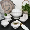 A collection of white ceramic soup tureens and bowls, some filled with food, arranged on a black surface. A decorative wooden tray with a heart design and a basket of parsley are in the background. Produktbild-Vorschau