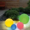 Four illuminated spherical garden lights in various colors are arranged on a wooden deck in a garden setting. Produktbild-Vorschau
