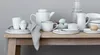 A collection of white ceramic tableware, including plates, cups, a teapot, and a pitcher, arranged on a wooden table. Produktbild-Vorschau