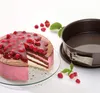 A round cake with pink frosting and raspberry topping sits on a glass plate next to a springform pan. Anteprima dell’immagine prodotto