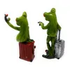 Two frog figurines, one taking a selfie with a phone and sitting on a red suitcase, the other standing with a silver suitcase. Produktbild-Vorschau