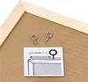 Close-up of two screw-in eye hooks and an instruction diagram on a brown backing board, with a wooden frame in the corner. Produktbild-Vorschau